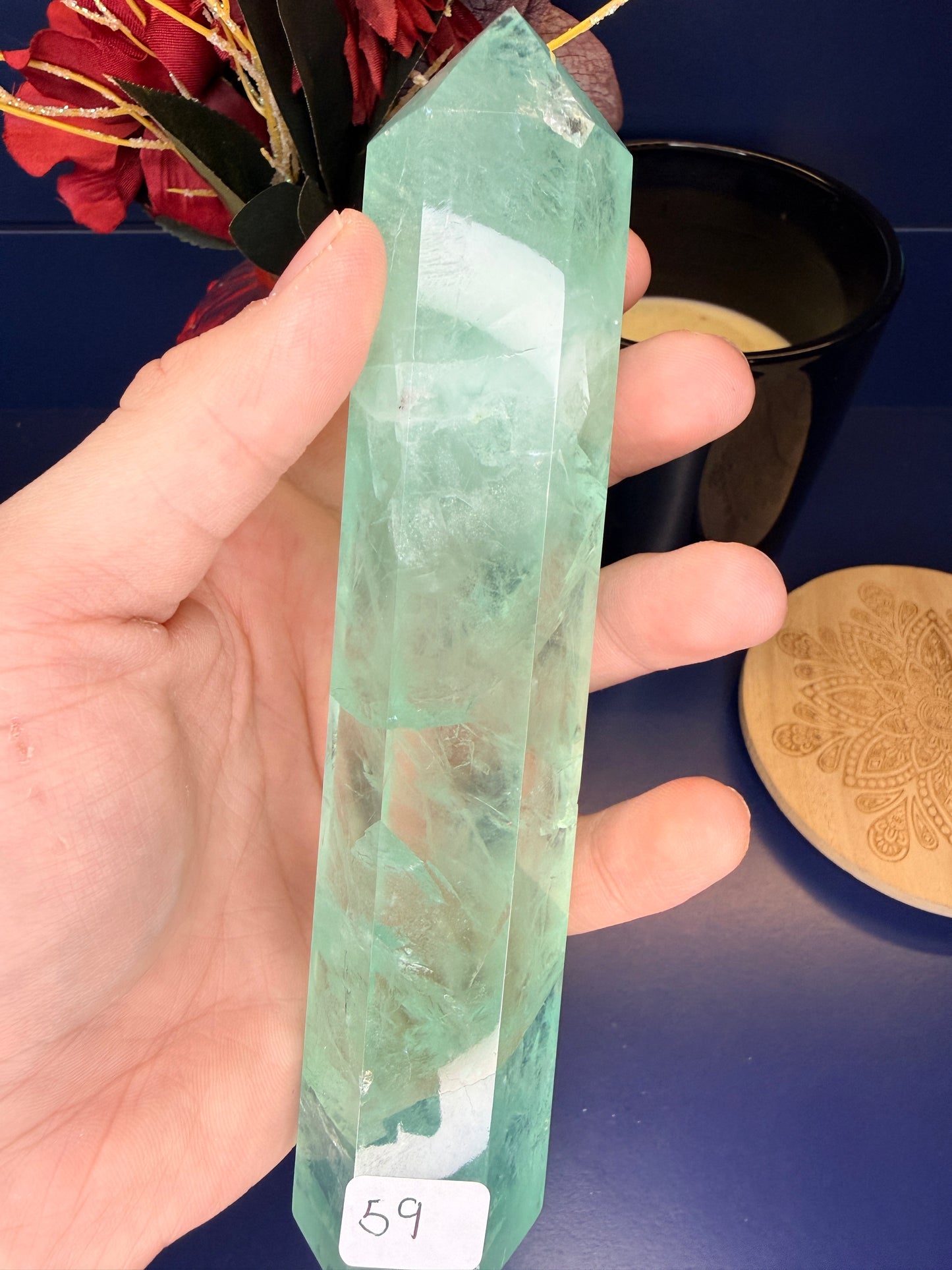 Hand holding a large translucent green fluorite tower in front of a red flower and black candle jar next to a wooden coaster