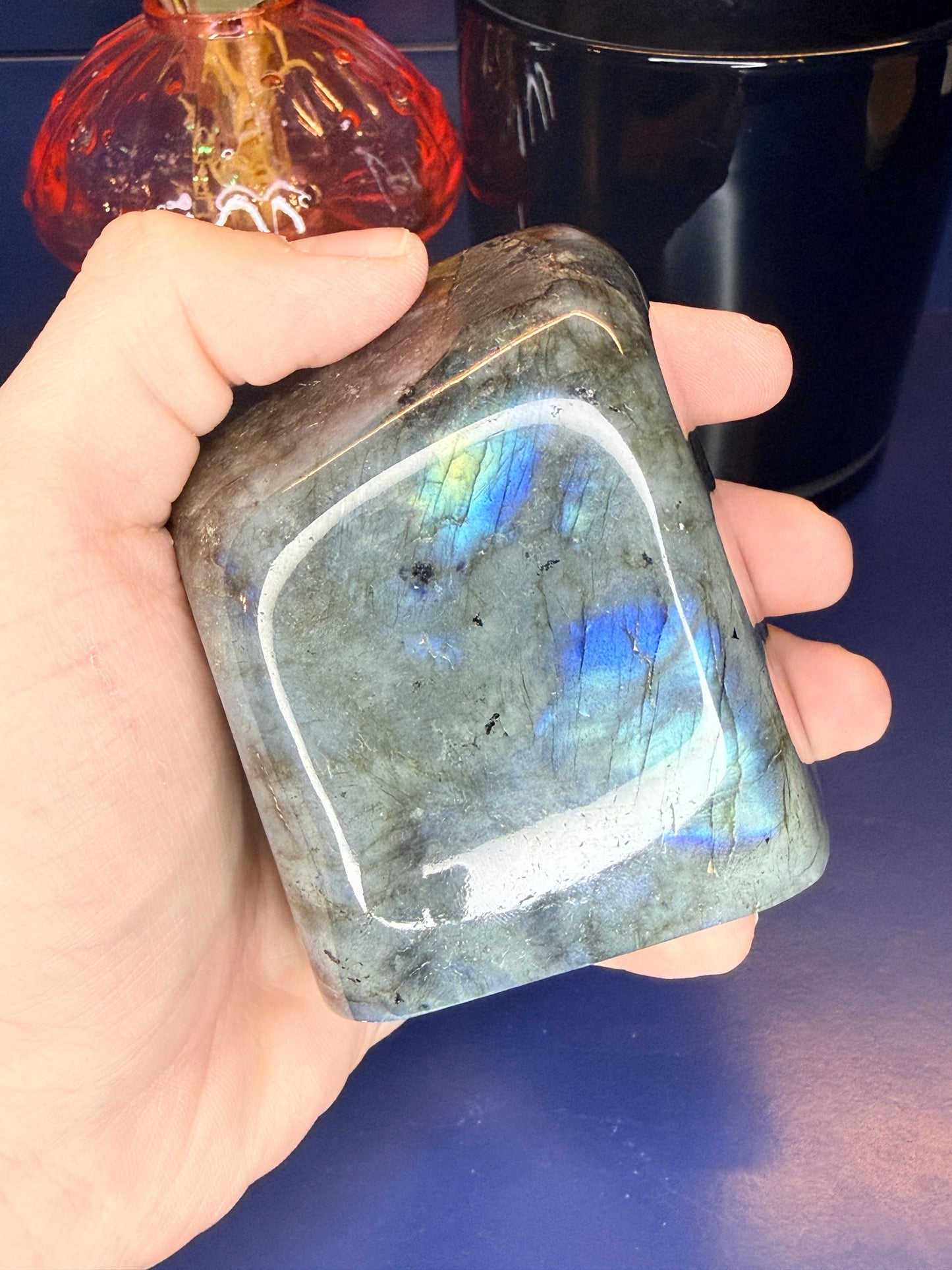 Hand holding a colorful blue flashing labradorite freeform in front of a mushroom vase and black jar