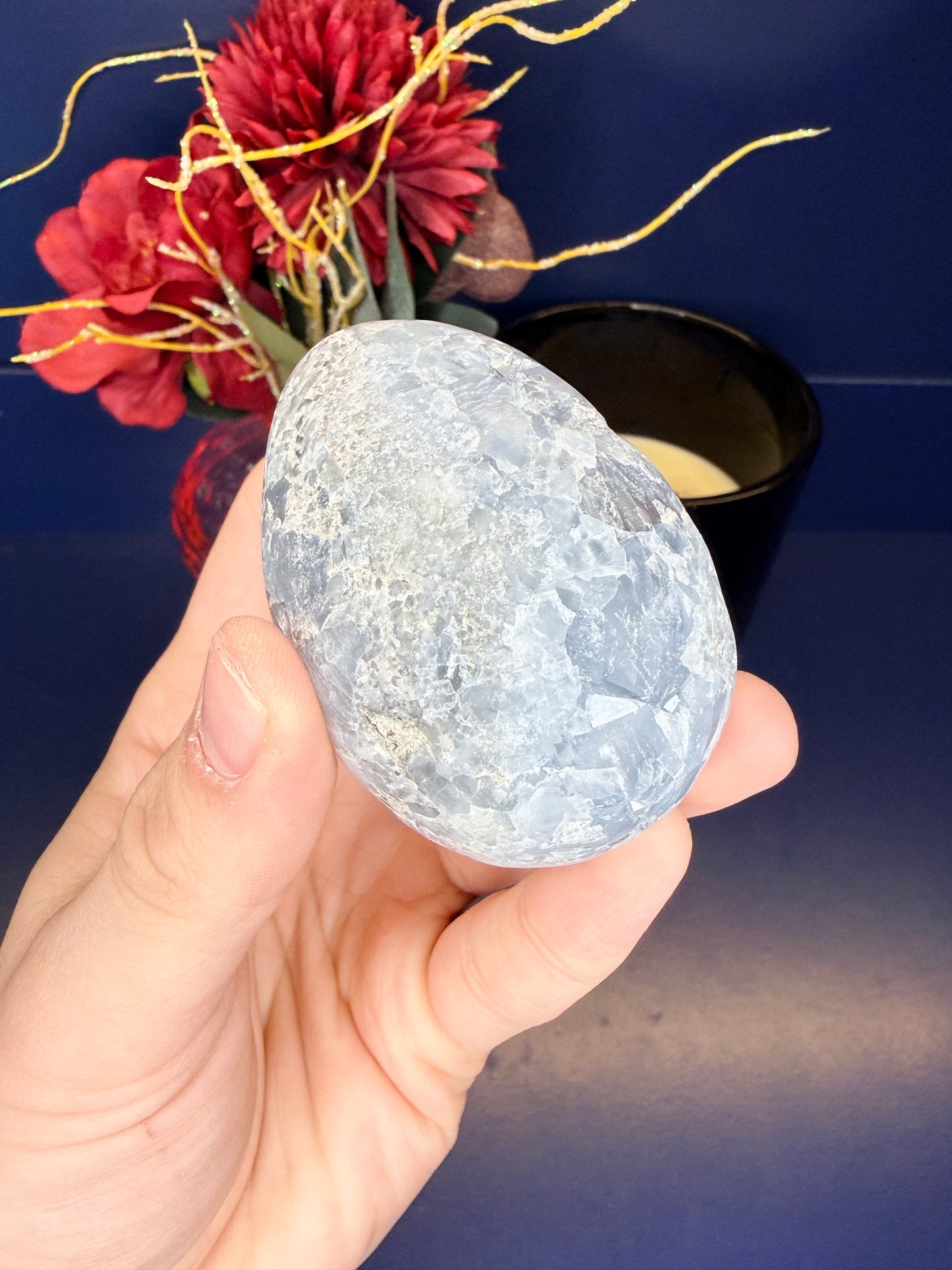Hand holding a blue and white celestite egg in front of red flowers and black jar on a blue background