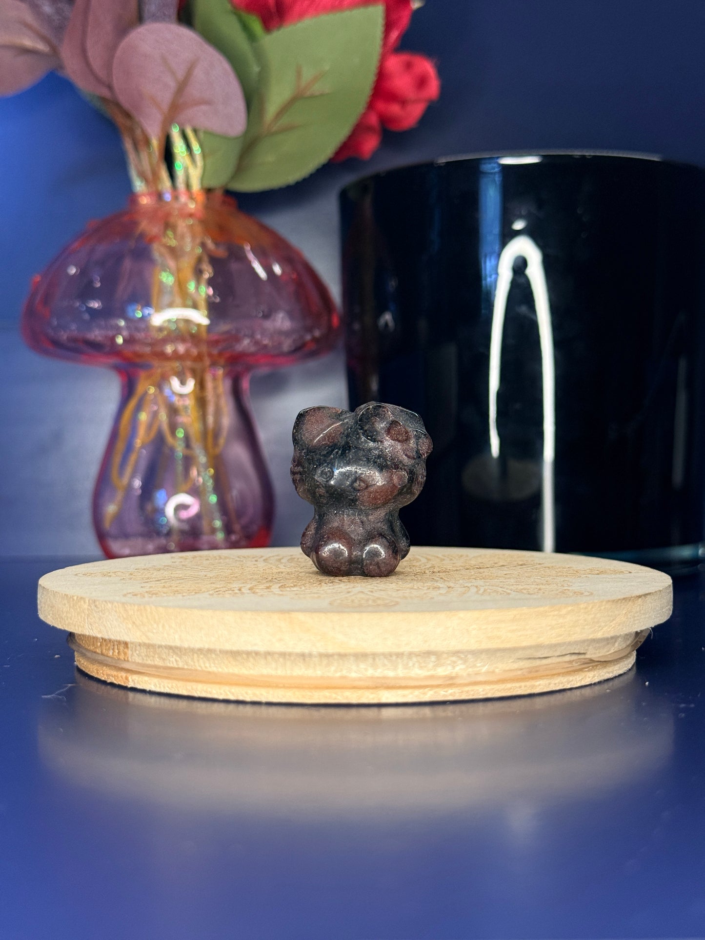 Red and black crystal kitty carving in front of a pink vase and black jar on a wooden coaster against a blue background