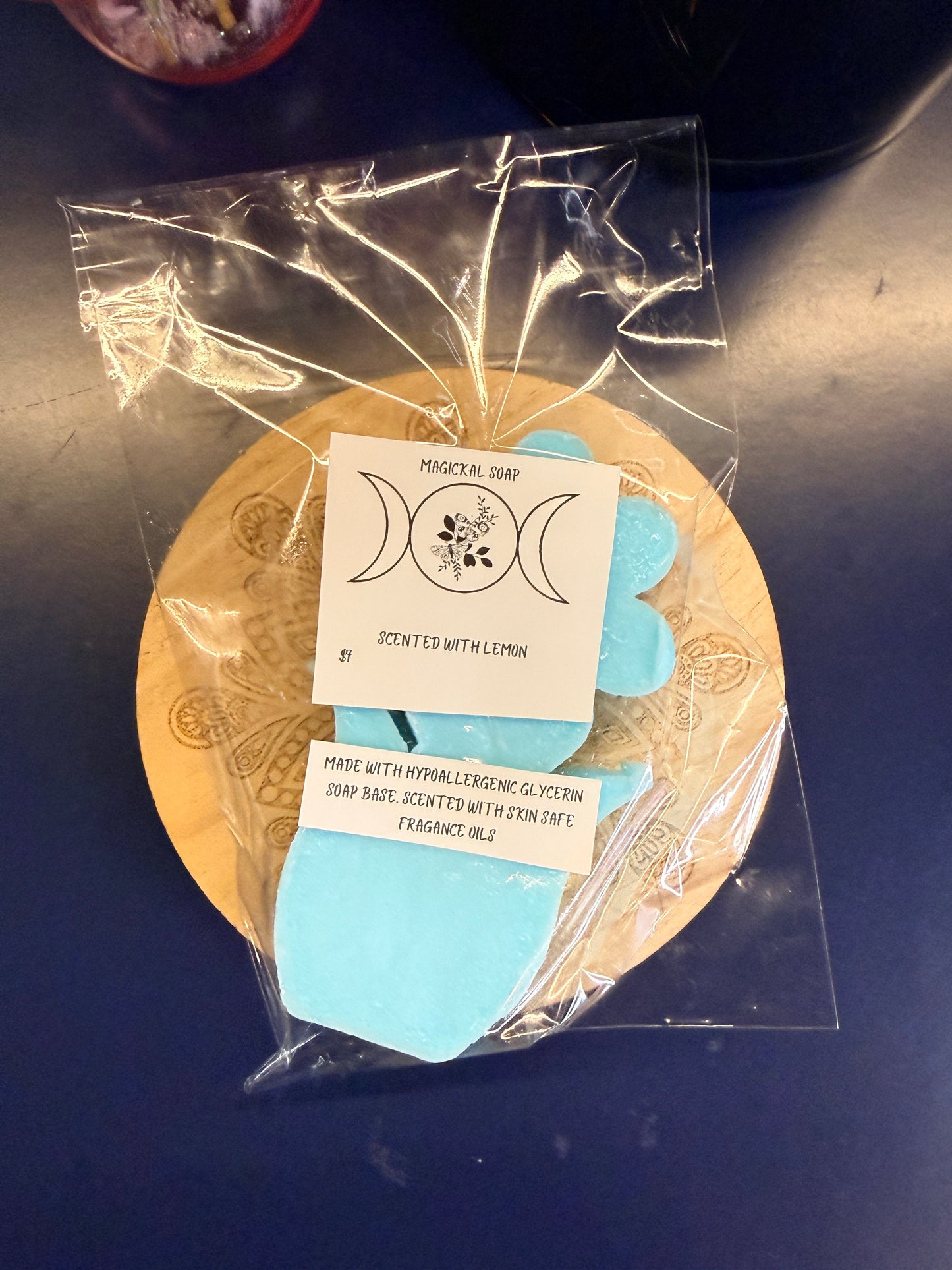 Blue and green flower soap bar with two labels on a wooden coaster, wrapped in clear plastic on a dark surface.