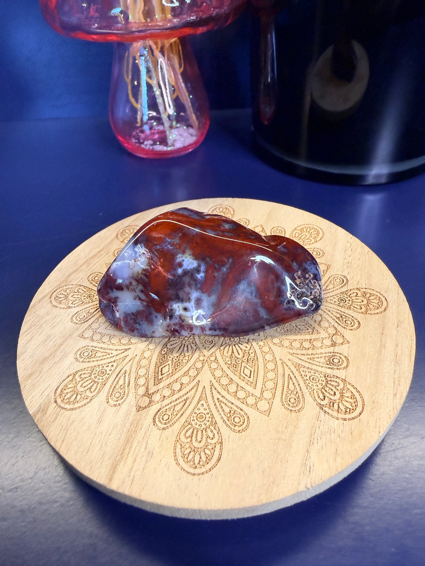 Red and light blue mexican agate freeform on a decorative wooden coaster in front of a mushroom and black jar