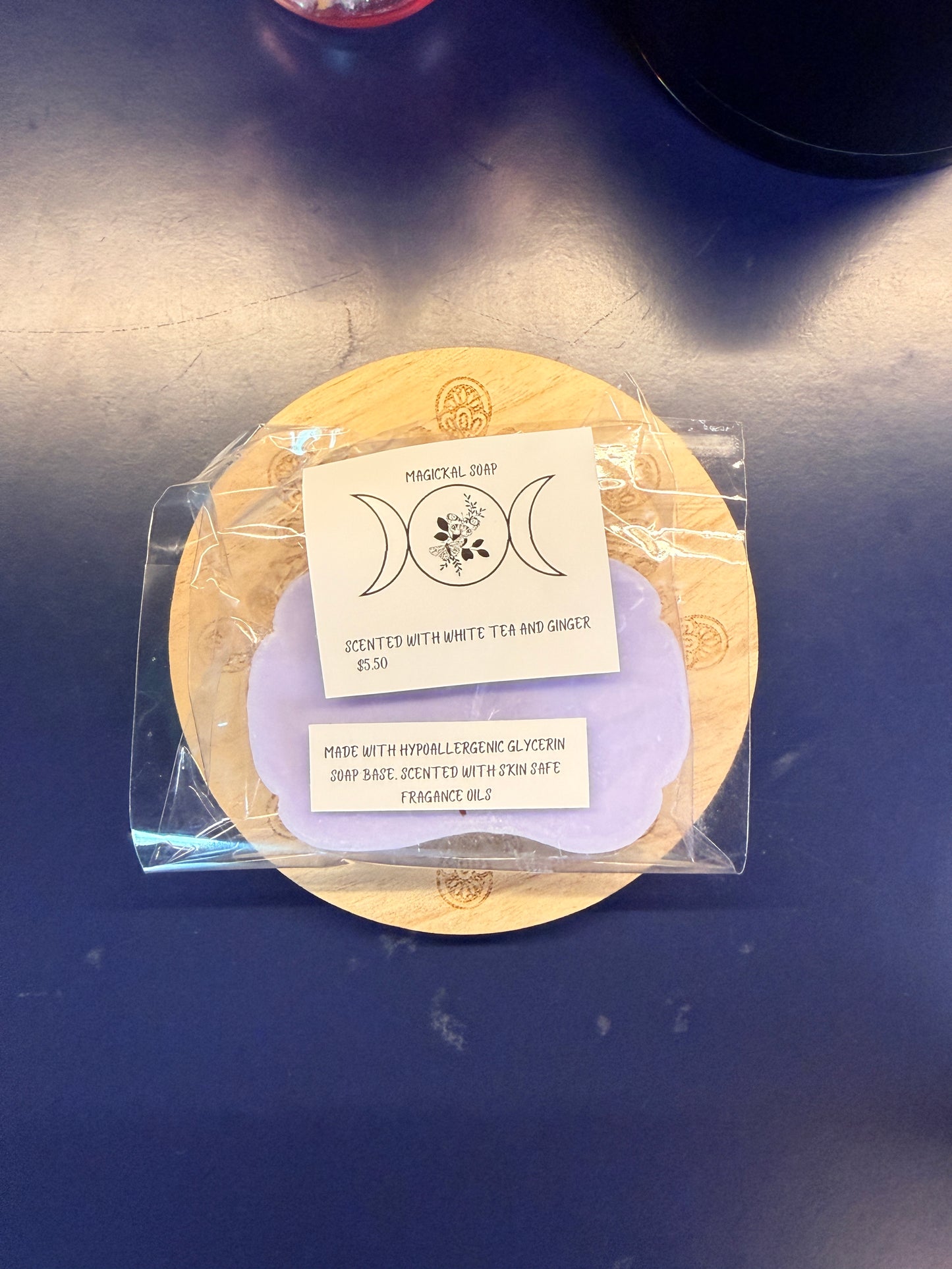 Purple soap bar with two labels on a wooden coaster, wrapped in clear plastic on a dark surface.