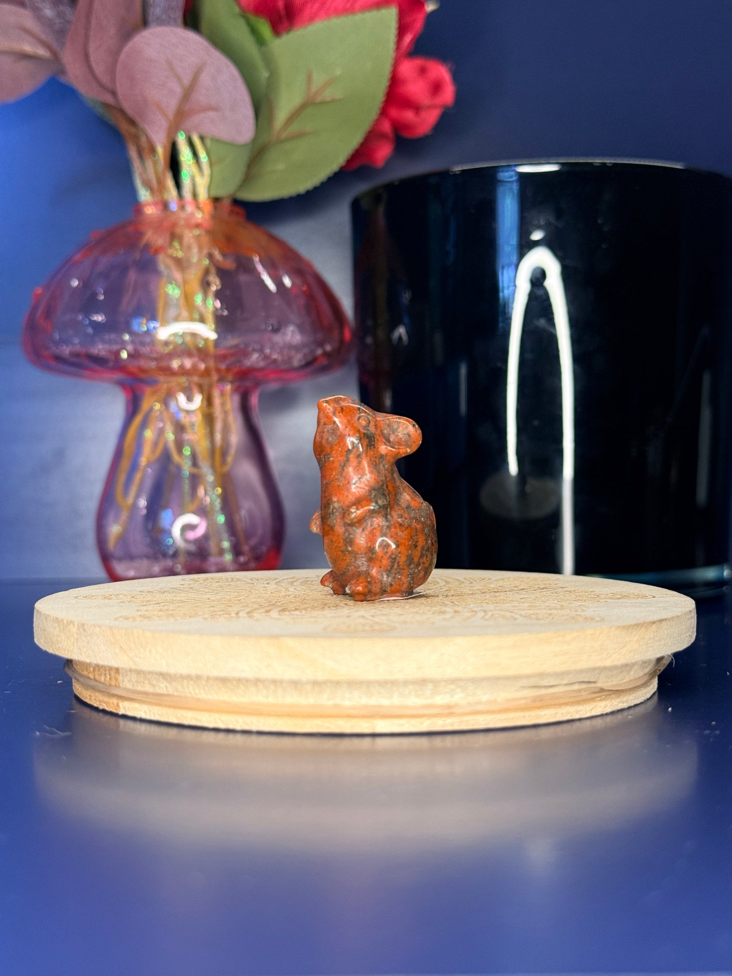 red sesame jasper mouse carving in front of a pink vase and black jar on a wooden coaster against a blue background