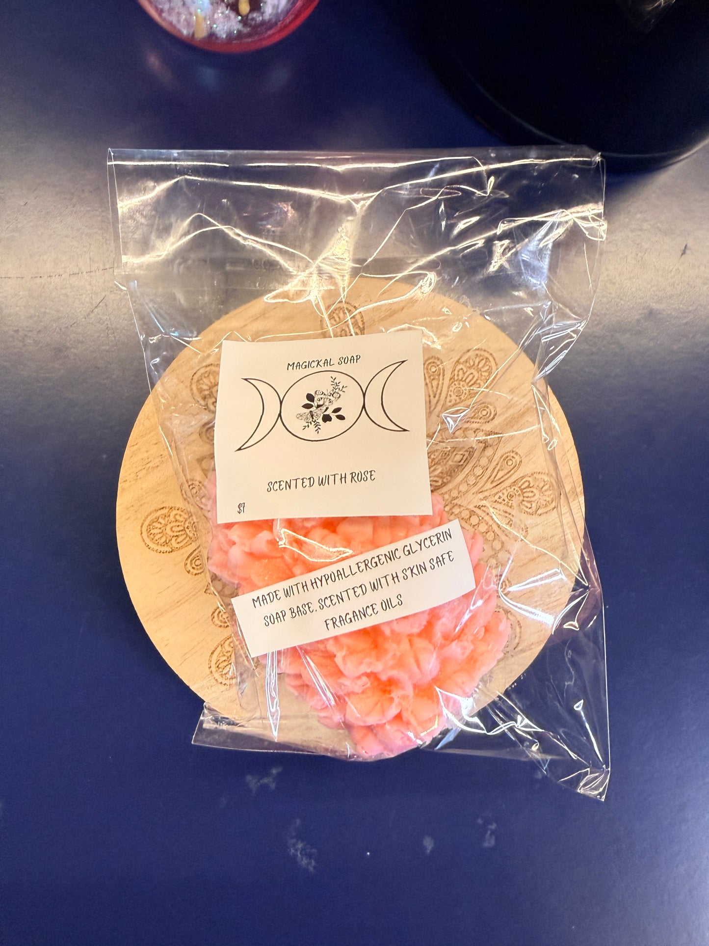 Orange flower-shaped soap with two labels on a decorative wooden coaster with clear plastic wrap.