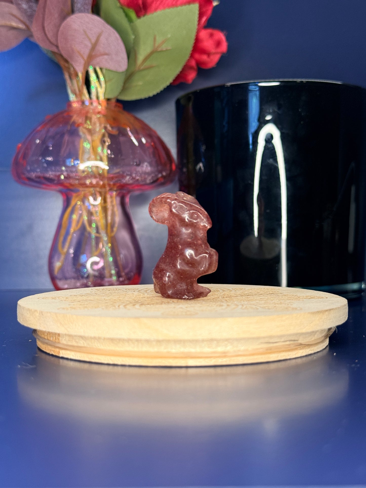 Pink strawberry quartz goat carving in front of a pink vase and black jar on a wooden coaster against a blue background