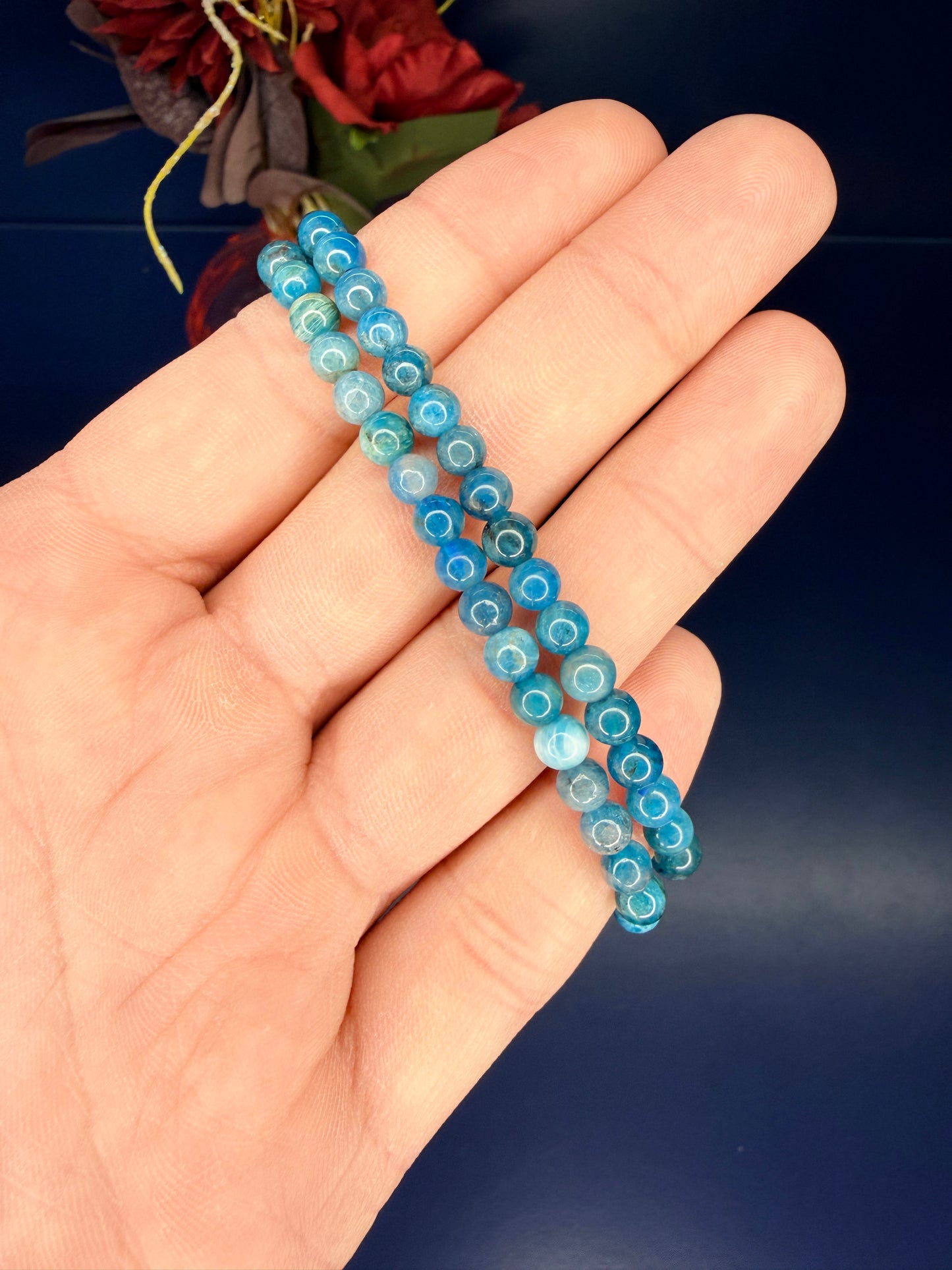 Hand holding two beaded blue blue apatite bracelets in front of a red flower on a blue background