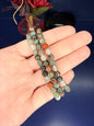 Hand holding three green and red beaded bloodstone bracelet in front of a red flower and blue background