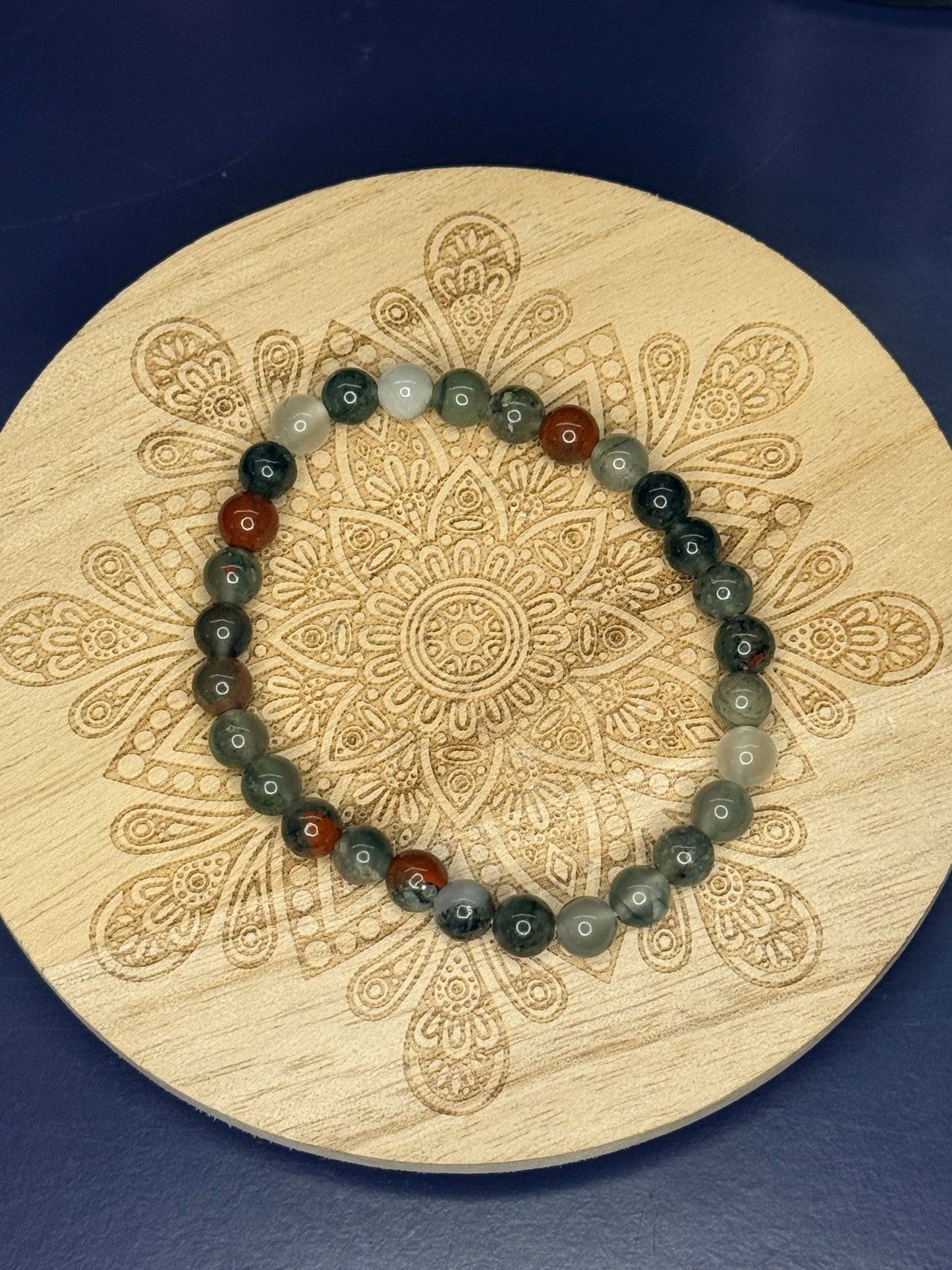 Green and red beaded bloodstone bracelet resting on a decorative wooden coaster on a blue background