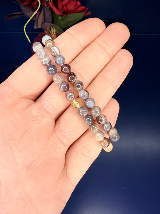 Hand holding two multicolored beaded botswana agate bracelets in front of a red flower and blue background