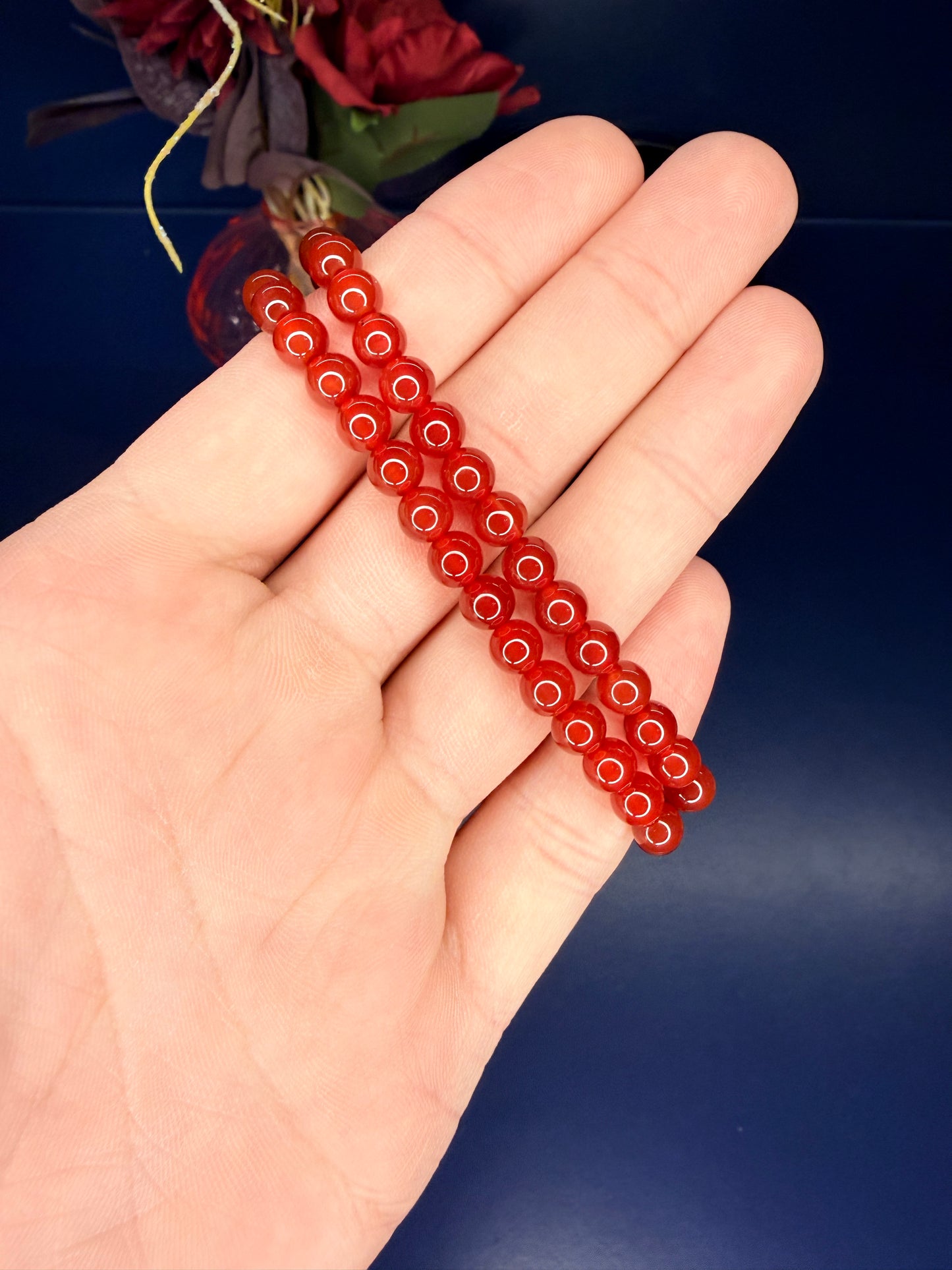 Hand holding two red beaded carnelian bracelets in front of a red flower on a blue background