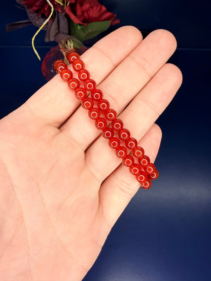 Hand holding two red beaded carnelian bracelets in front of a red flower on a blue background
