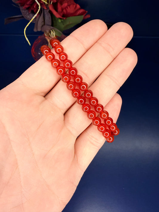 Hand holding two red beaded carnelian bracelets in front of a red flower on a blue background