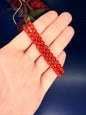 Hand holding two red beaded carnelian bracelets in front of a red flower on a blue background