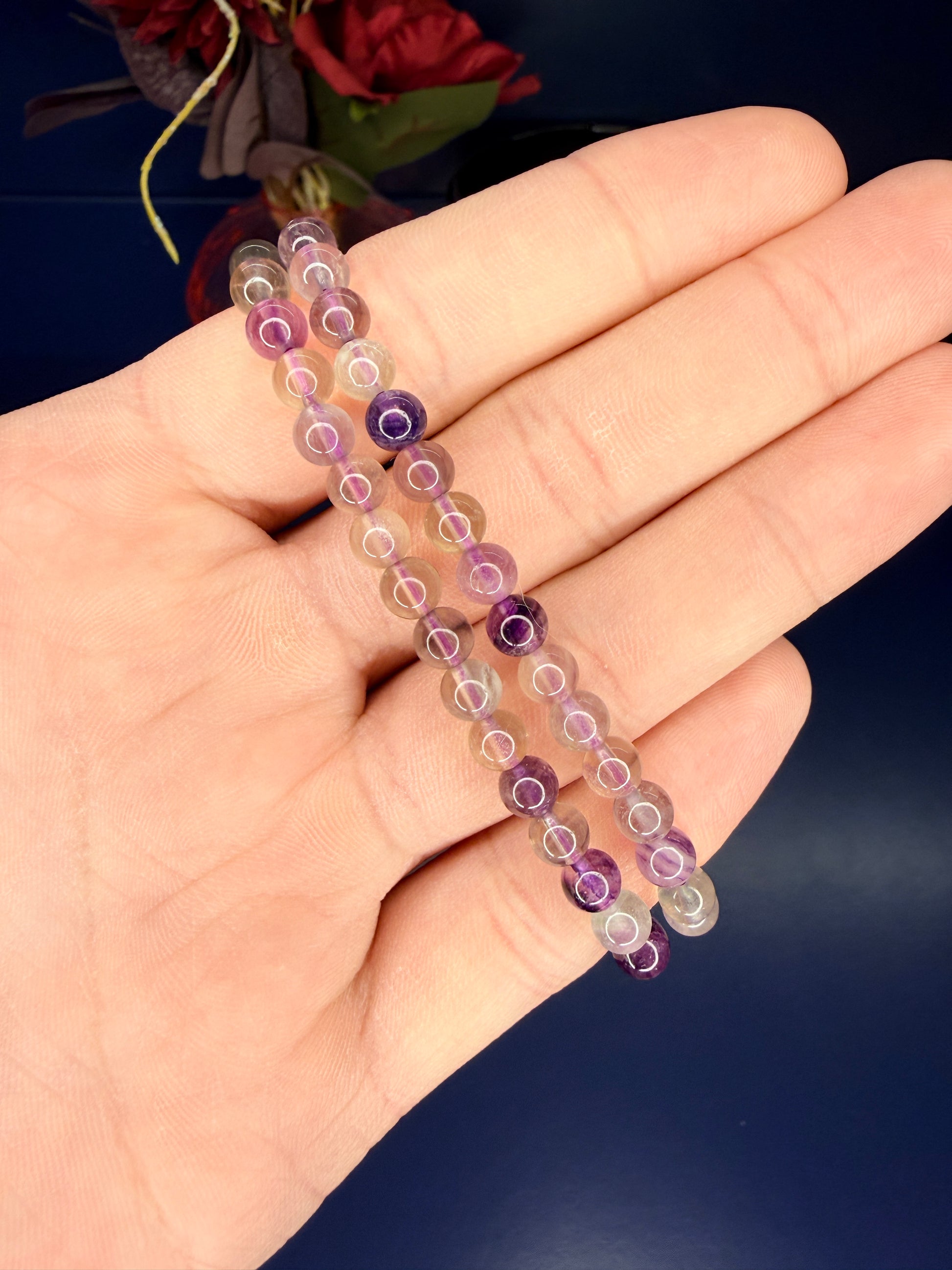 Hand holding two translucent beaded fluorite bracelets in front of a red flower and blue background