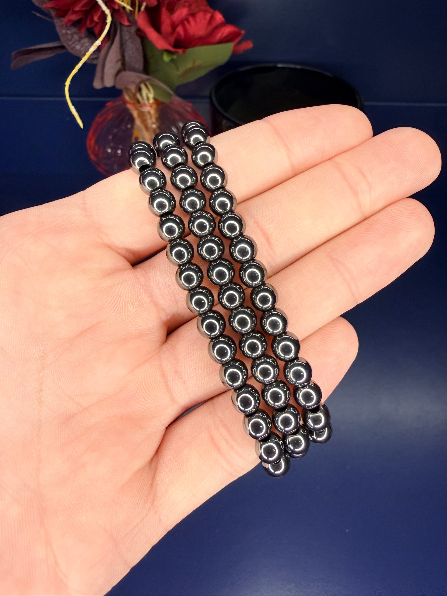 Hand holding three shining black beaded hematite bracelets in front of a red flower and blue background