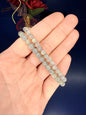 Hand holding two grey beaded labradorite bracelet in front of a red flower and blue background