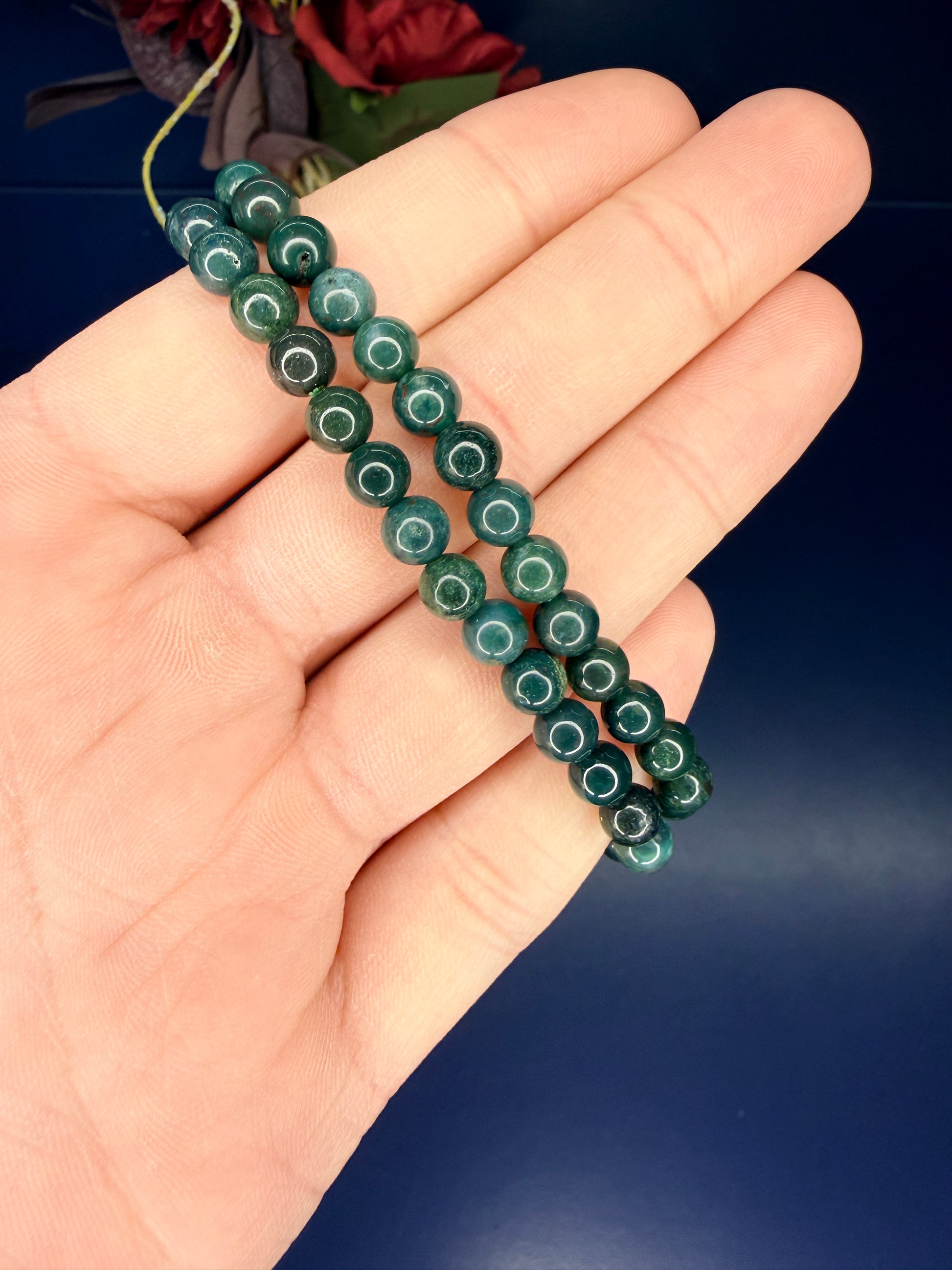 Hand holding two green beaded moss agate bracelets in front of a red flower on a blue background