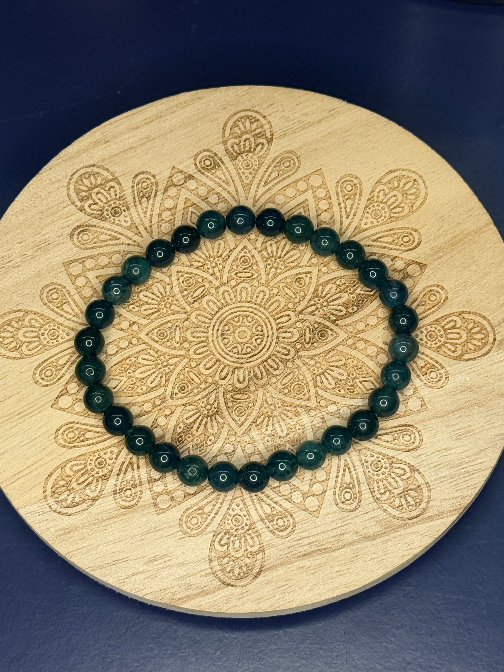 Green beaded moss agate bracelet resting on a decorative wooden coaster on a blue background