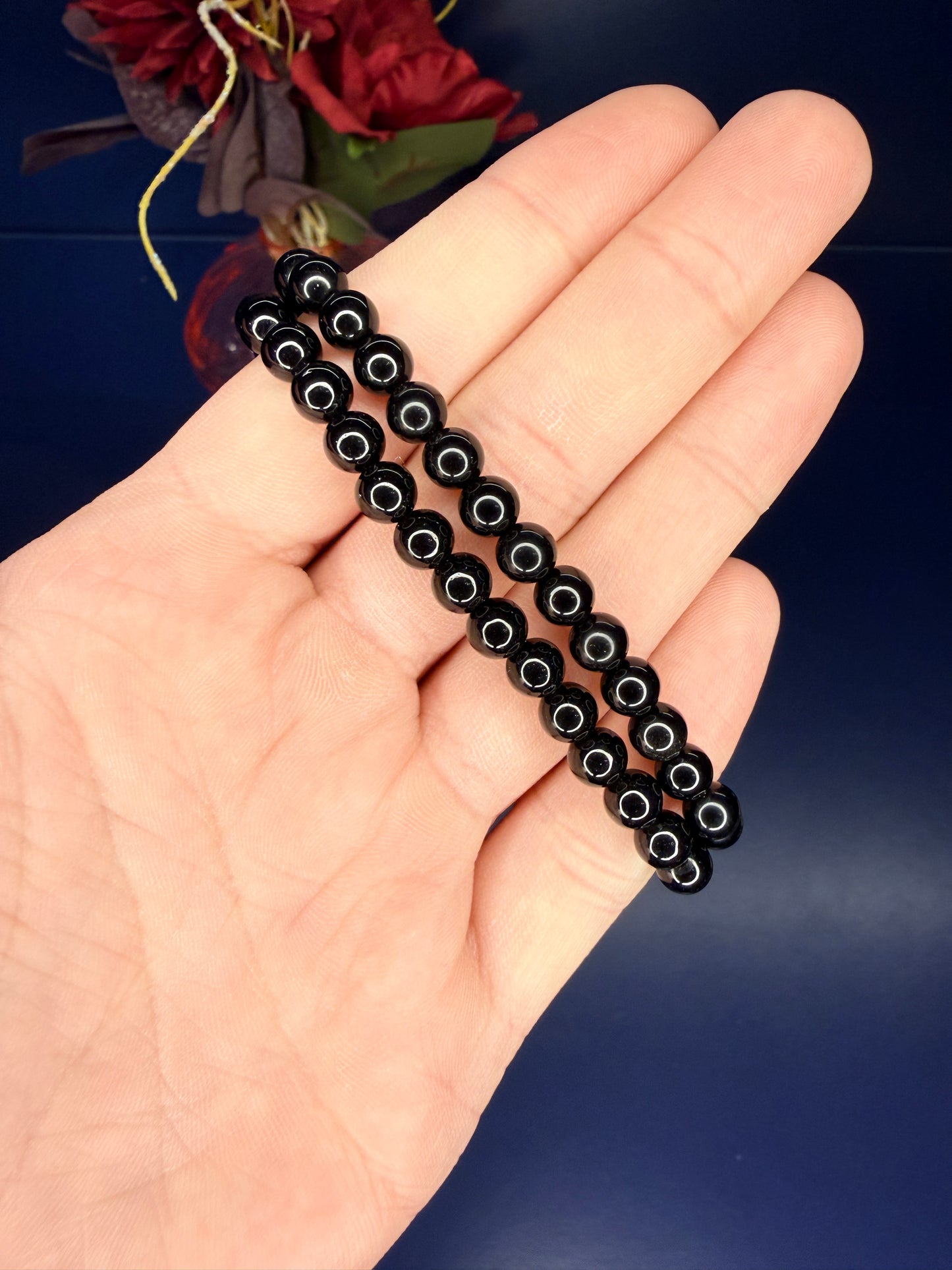 Hand holding two black beaded obsidian bracelets in front of a red flower on a blue background