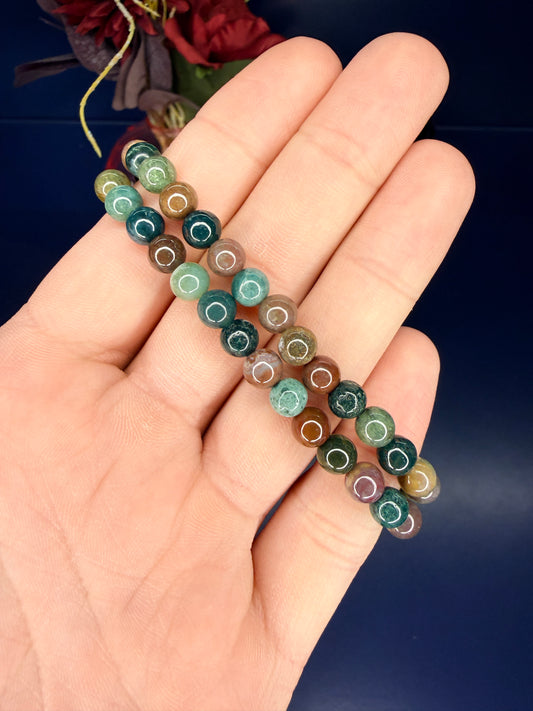 Hand holding two multicolored beaded ocean jasper bracelets in front of a red flower and blue background