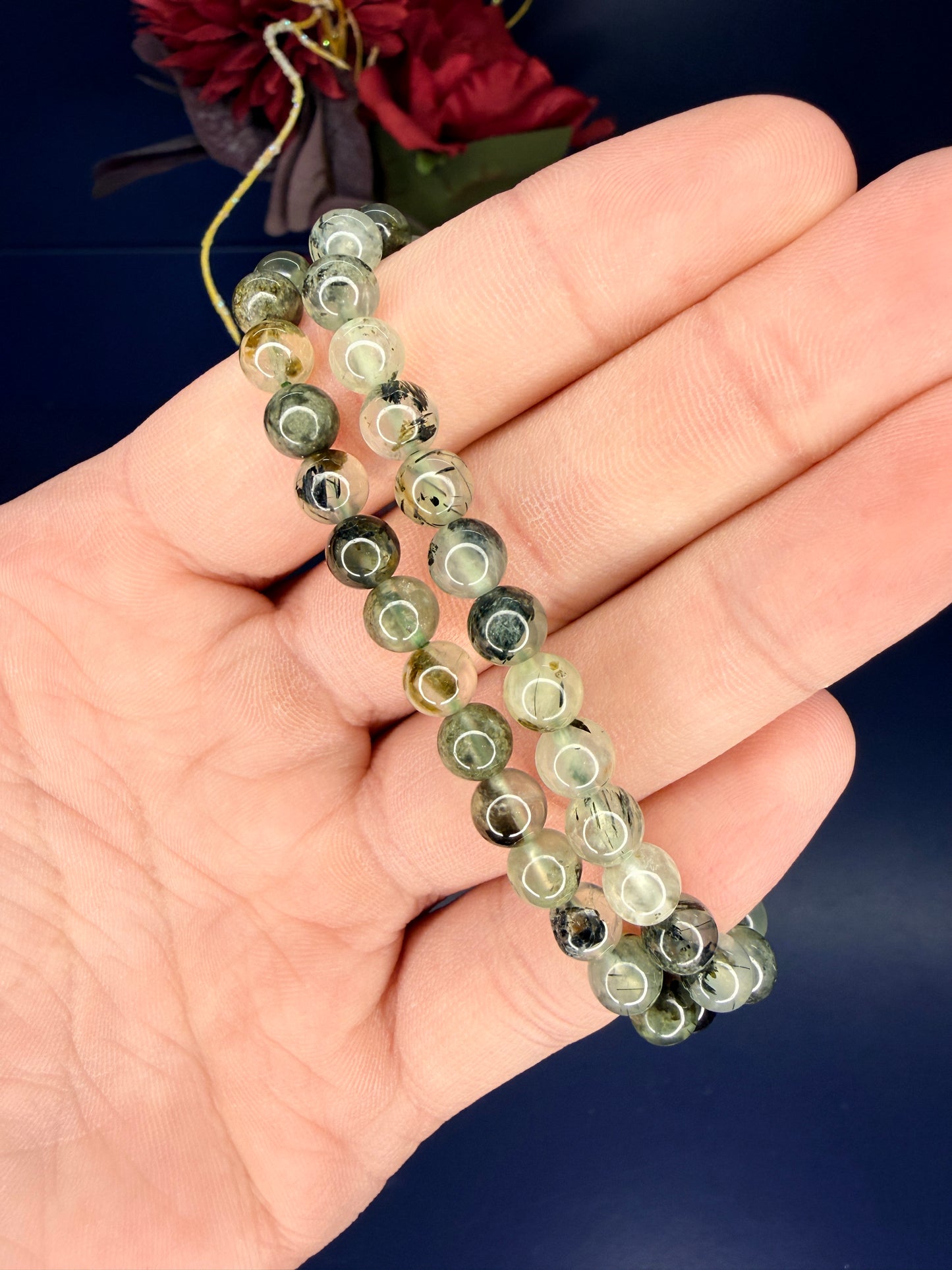 Hand holding two beaded green prehnite bracelets in front of a red flower on a blue background