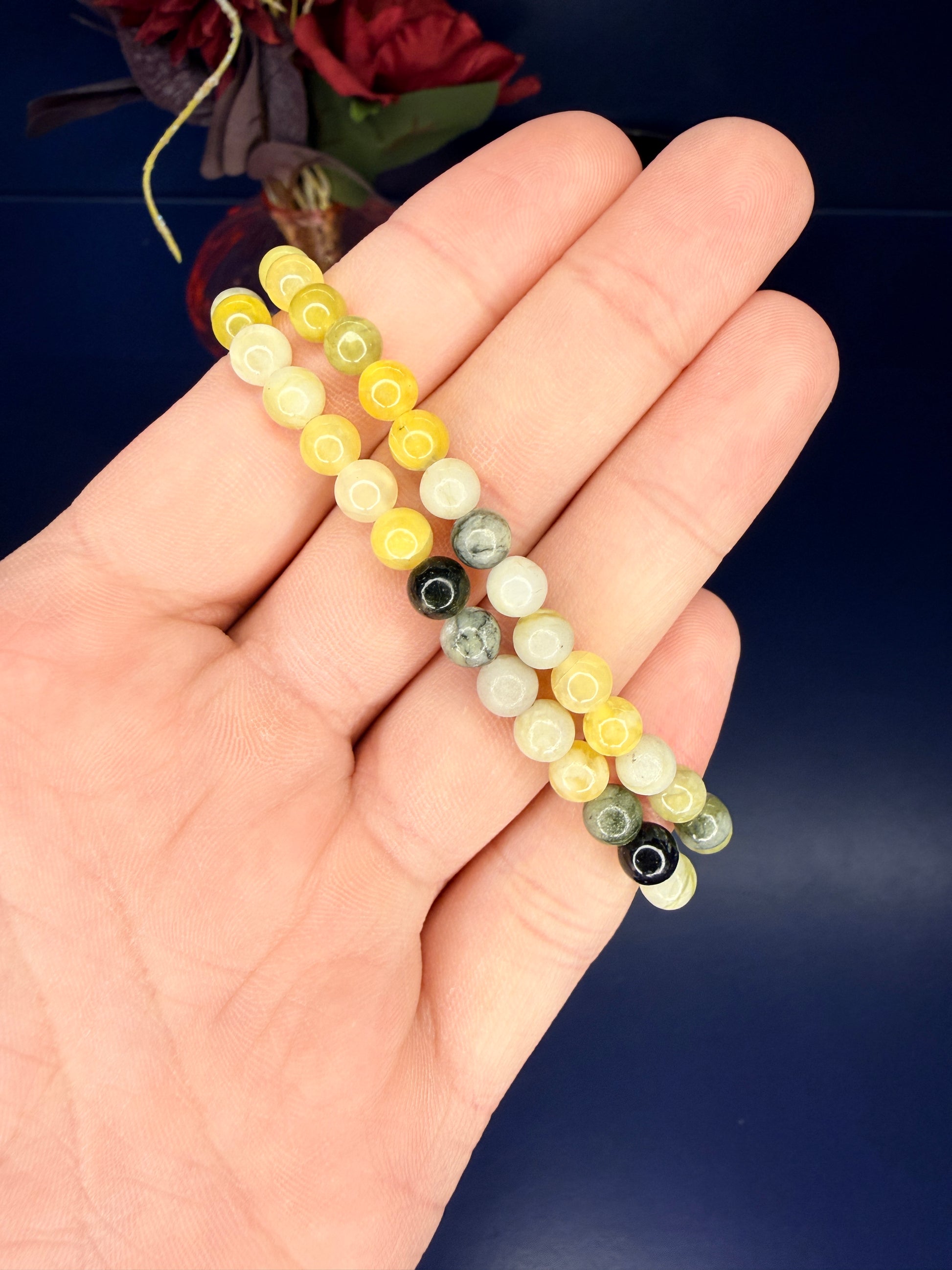 Hand holding two green and yellow beaded serpentine bracelets in front of a red flower on a blue background