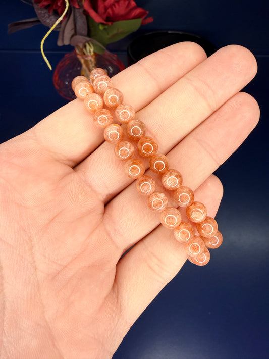 Hand holding two beaded orange sunstone bracelets in front of a red flower on a blue background