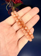 Hand holding two beaded orange sunstone bracelets in front of a red flower on a blue background