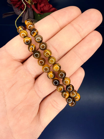 Hand holding two golden beaded tiger's eye bracelets in front of a red flower and blue background