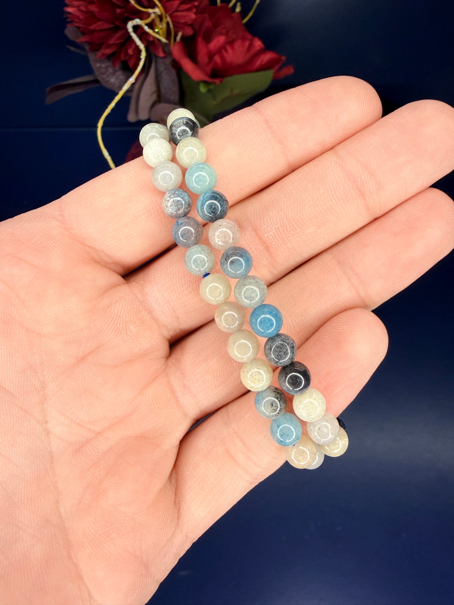 Hand holding two blue and beige beaded trolleite bracelets in front of a red flower and blue background
