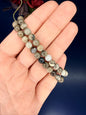 Hand holding two multicolored beaded volcano agate bracelets in front of a red flower on a blue background