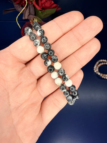 Hand holding two black and white beaded zebra jasper bracelets in front of a red flower on a blue background