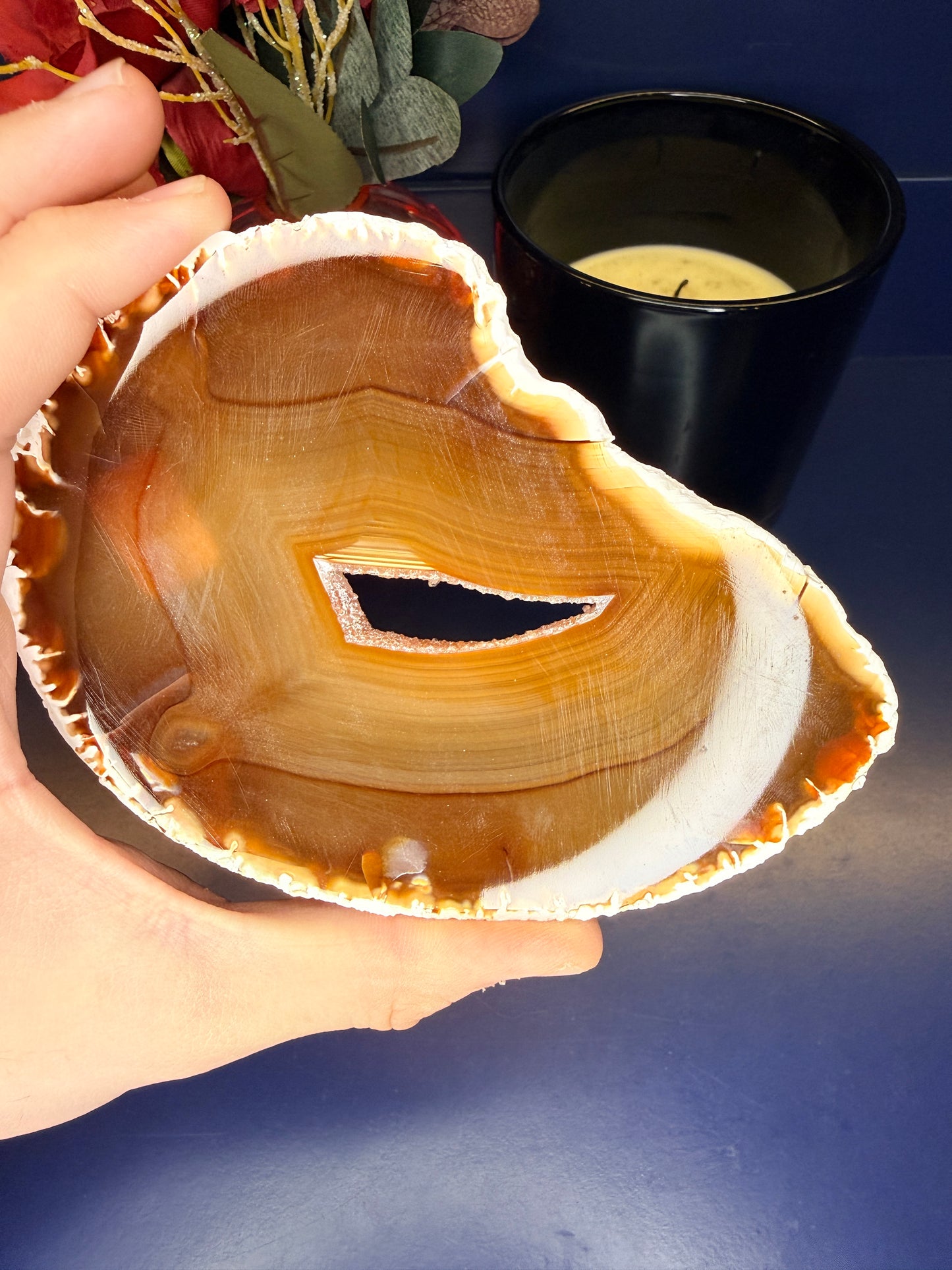 Hand holding an amber colored agate slice with a druzy in the middle in front of a red flower and black candle jar