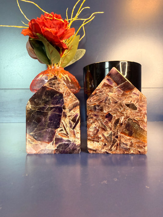 Two Purple and amber alien amethyst freeforms with intricate patterns sitting in front of a red flower and black jar