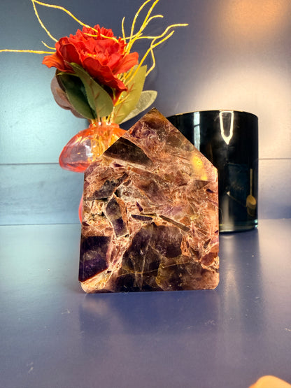 A purple and amber alien amethyst freeforms with intricate patterns sitting in front of a red flower and black jar