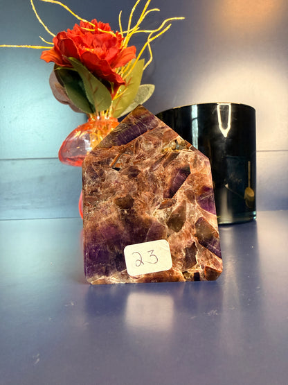 A purple and amber alien amethyst freeforms with intricate patterns sitting in front of a red flower and black jar