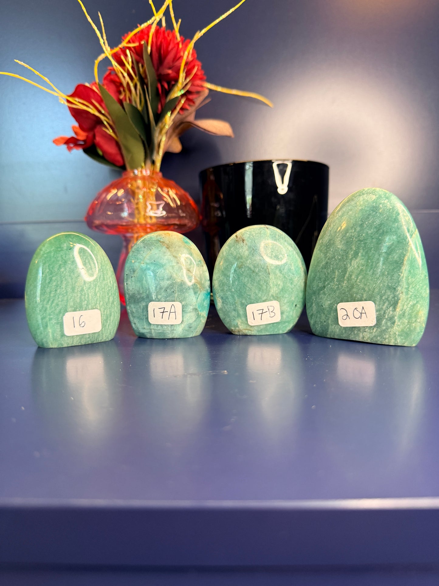 Four blue green oblong amazonite free forms in front of a red flower in a vase and a black jar on a blue background