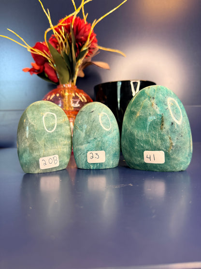 Three blue green oblong amazonite free forms in front of a red flower in a vase and a black jar on a blue background