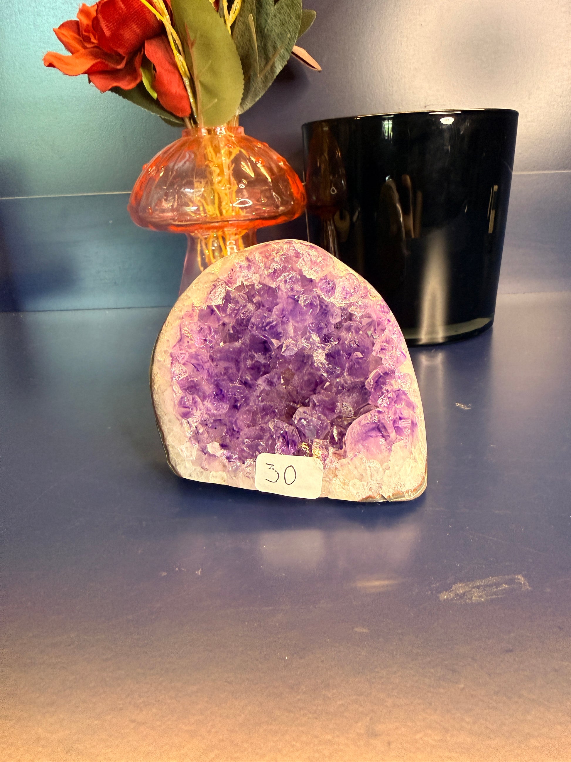 Bright purple and white amethyst cathedral in front of a red flower in a vase and black jar on a blue background