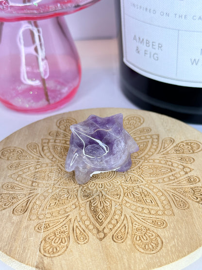 A purple Amethyst gengar carving resting on a wooden coaster in front of a black jar and pink vase on a white background