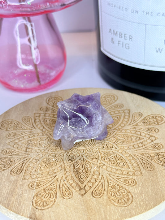 A purple Amethyst gengar carving resting on a wooden coaster in front of a black jar and pink vase on a white background