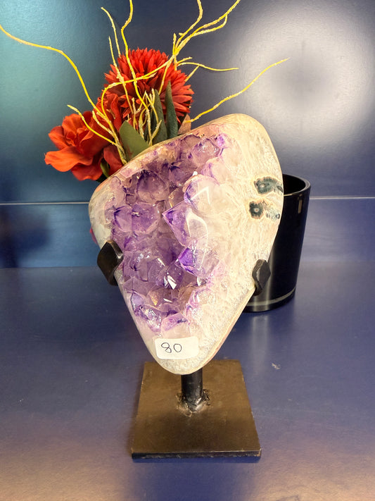 Large purple and white amethyst geode on a black metal stand in front of a red flower on a blue background