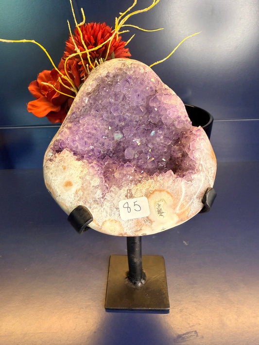Large purple and white amethyst geode on a black metal stand in front of a red flower on a blue background
