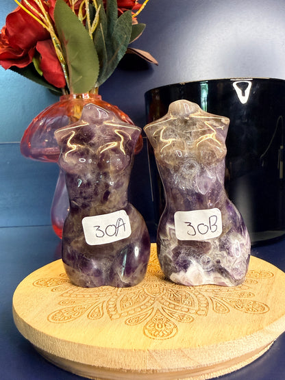 Two deep purple amethyst goddess bodies on a wooden coaster in front of a red flower in a vase and black jar