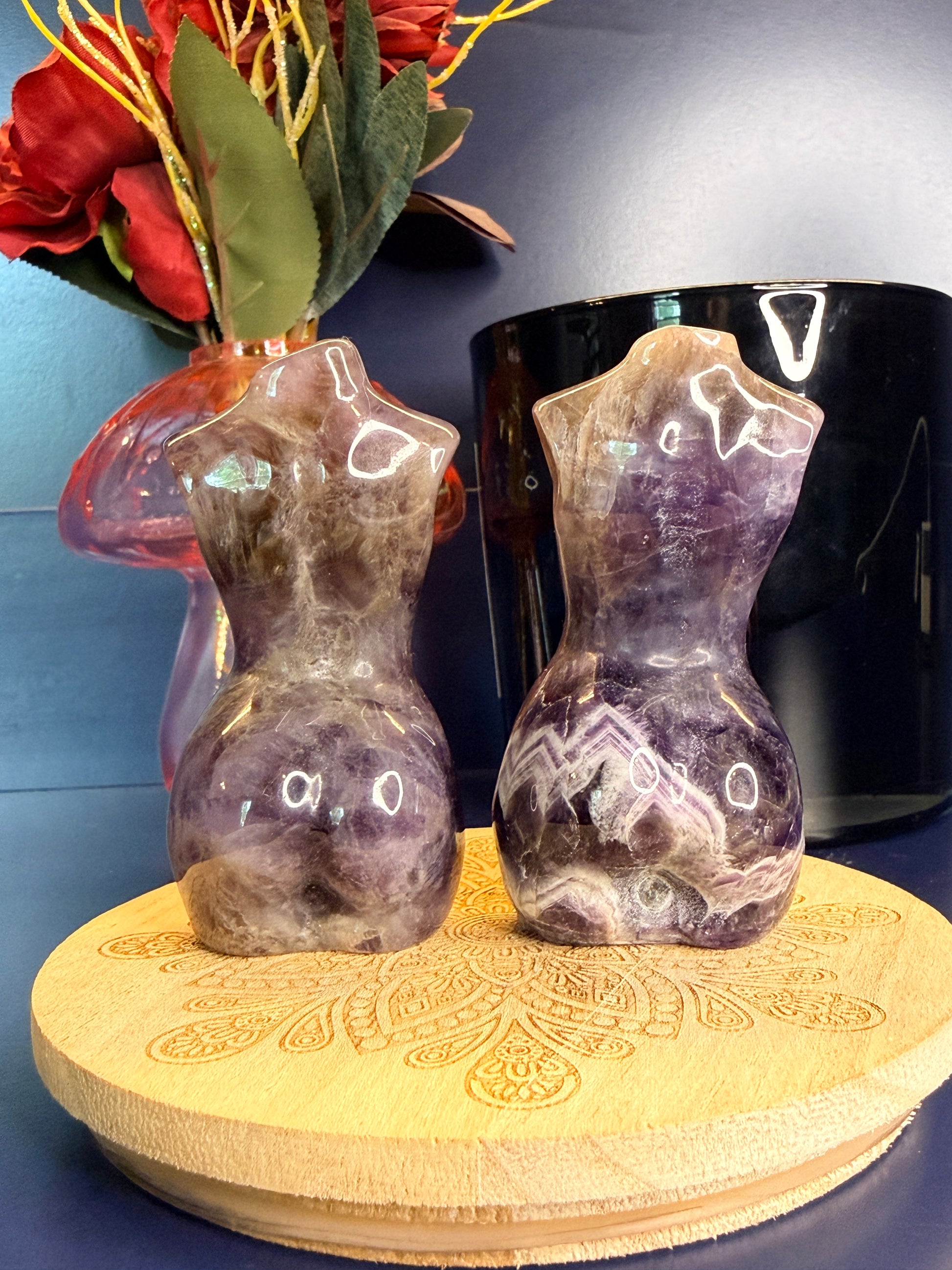Two deep purple amethyst goddess bodies on a wooden coaster in front of a red flower in a vase and black jar