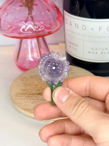 Hand holding a purple and green amethyst sunflower carving in front of a black jar and pink vase