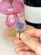 Hand holding a purple and green amethyst sunflower carving in front of a black jar and pink vase
