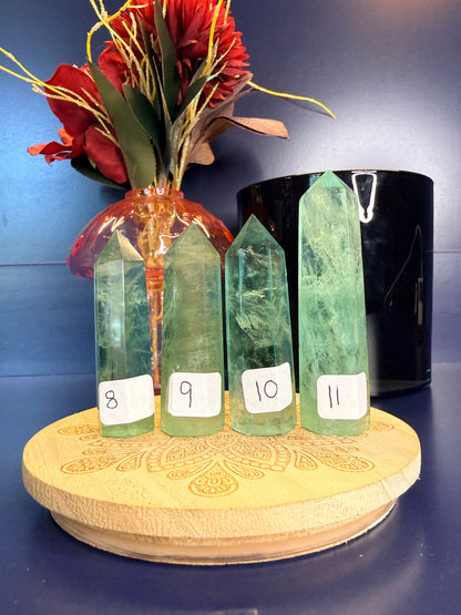 Four translucent green fluorite towers on a wooden coaster in front of a red flower and black jar on a blue background