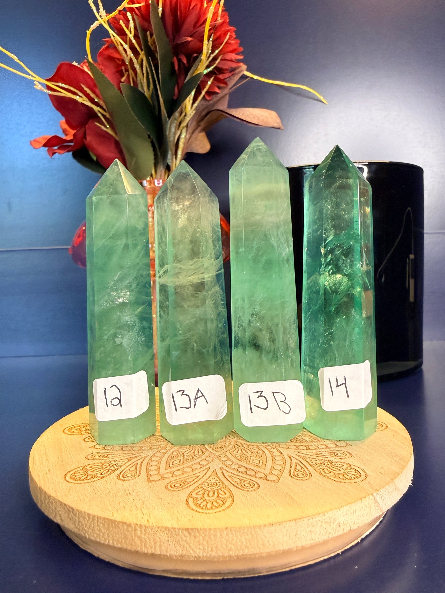 Four translucent green fluorite towers on a wooden coaster in front of a red flower and black jar on a blue background